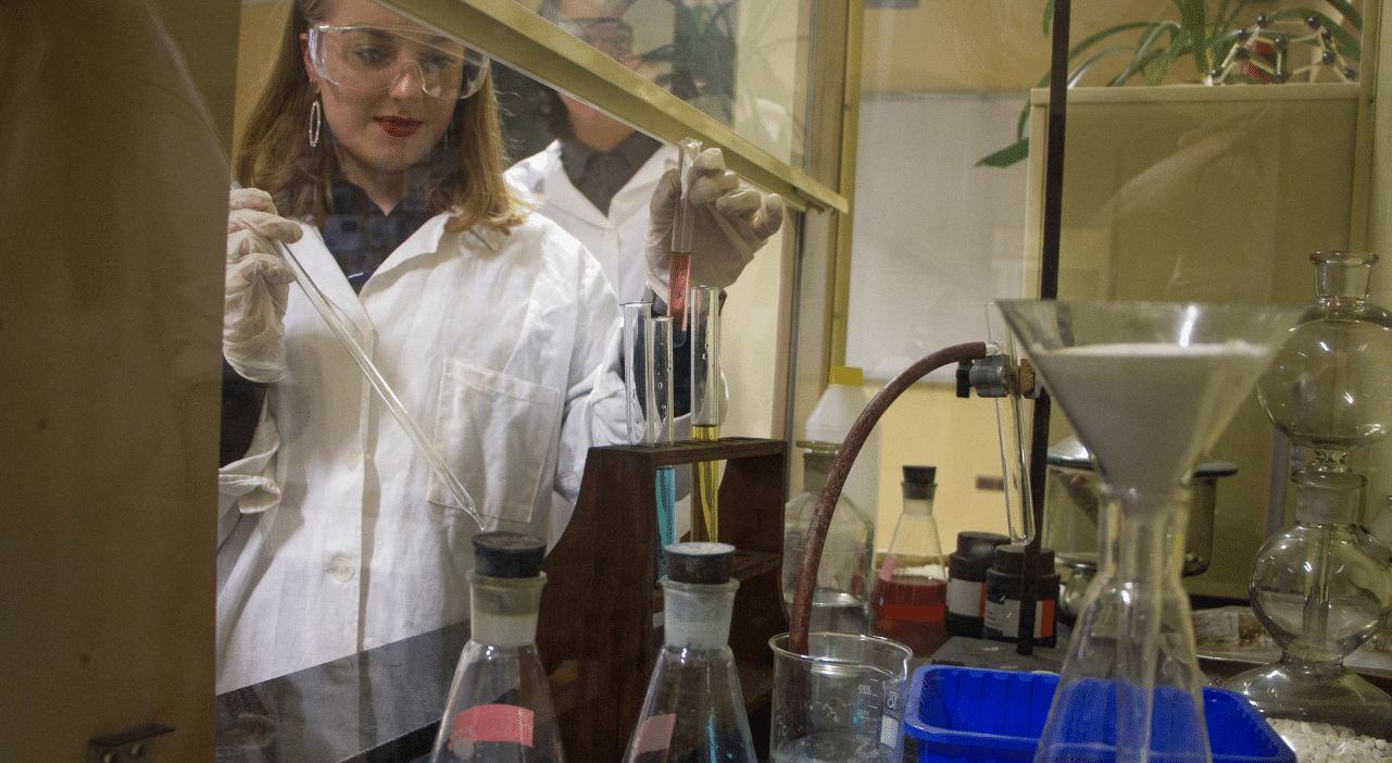 Zdjęcie przedstawiające uczniów w trakcie zajęć w pracowni chemicznej. Uczennica w fartuchu i okularach ochronnych pobiera pipetką substancję chemiczną ze szklanego pojemnika.